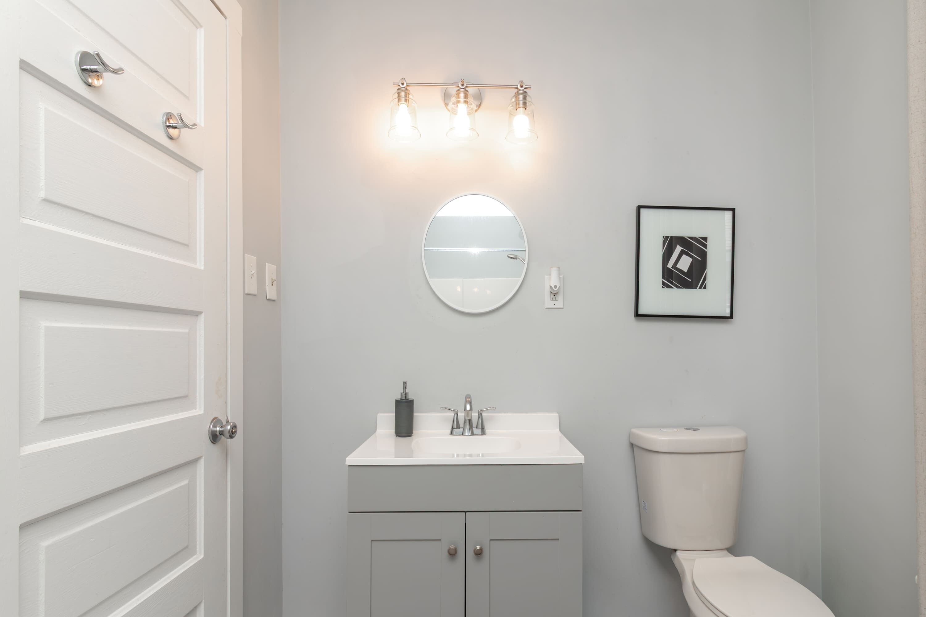 A bathroom with a white door, a round mirror, a sink with a faucet, a toilet, and a framed picture on the wall.