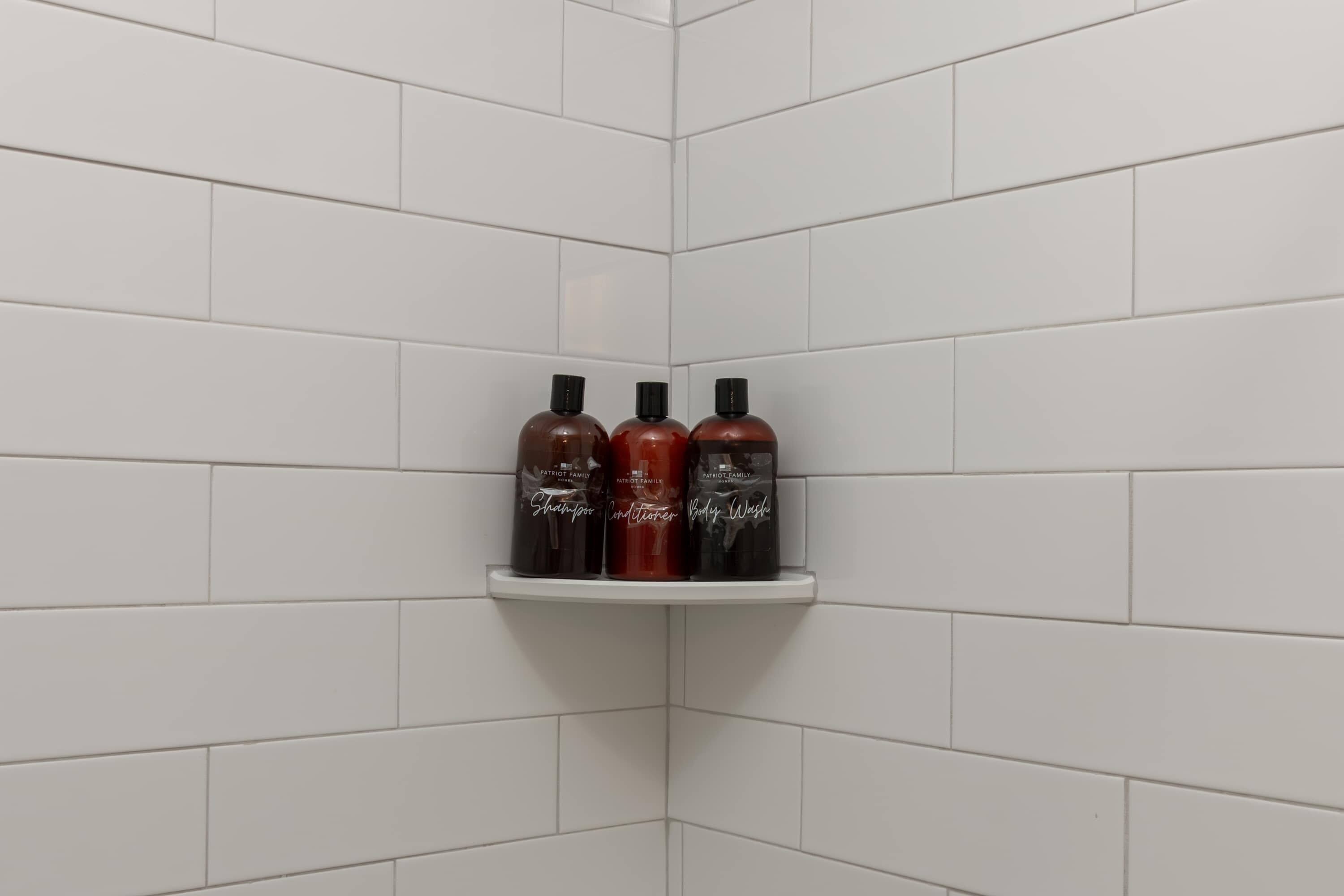 Angolo di un bagno con pareti rivestite di piastrelle bianche e uno scaffale bianco con tre flaconi di shampoo.