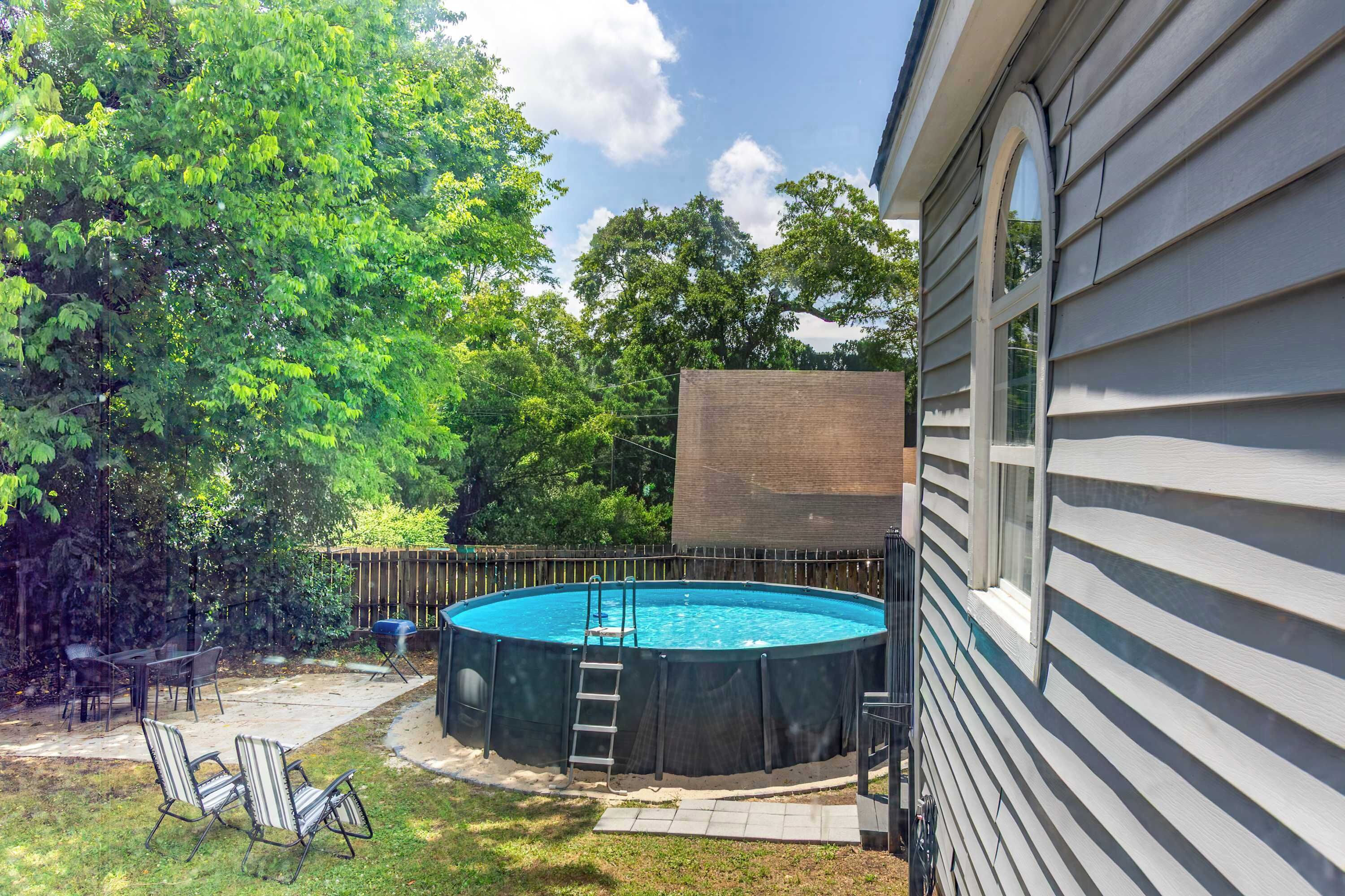 Una piscina exterior con escalera, rodeada por una terraza de madera y sillas, con árboles y una casa al fondo.