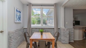 Ein Essbereich mit Holztisch, Stühlen, einer Topfpflanze, einem Fenster mit Vorhängen und einer Küche im Hintergrund.