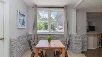 A dining area with a wooden table, chairs, a potted plant, a window with curtains, and a kitchen in the background.