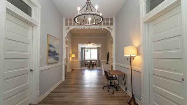 Ein Korridor mit Holzboden, weißen Türen, einem Chandelier, einem Essbereich mit Tisch und Stühlen sowie einer Stehlampe.