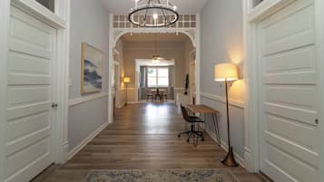 A hallway with wooden flooring, white doors, a chandelier, a dining area with a table and chairs, and a floor lamp.