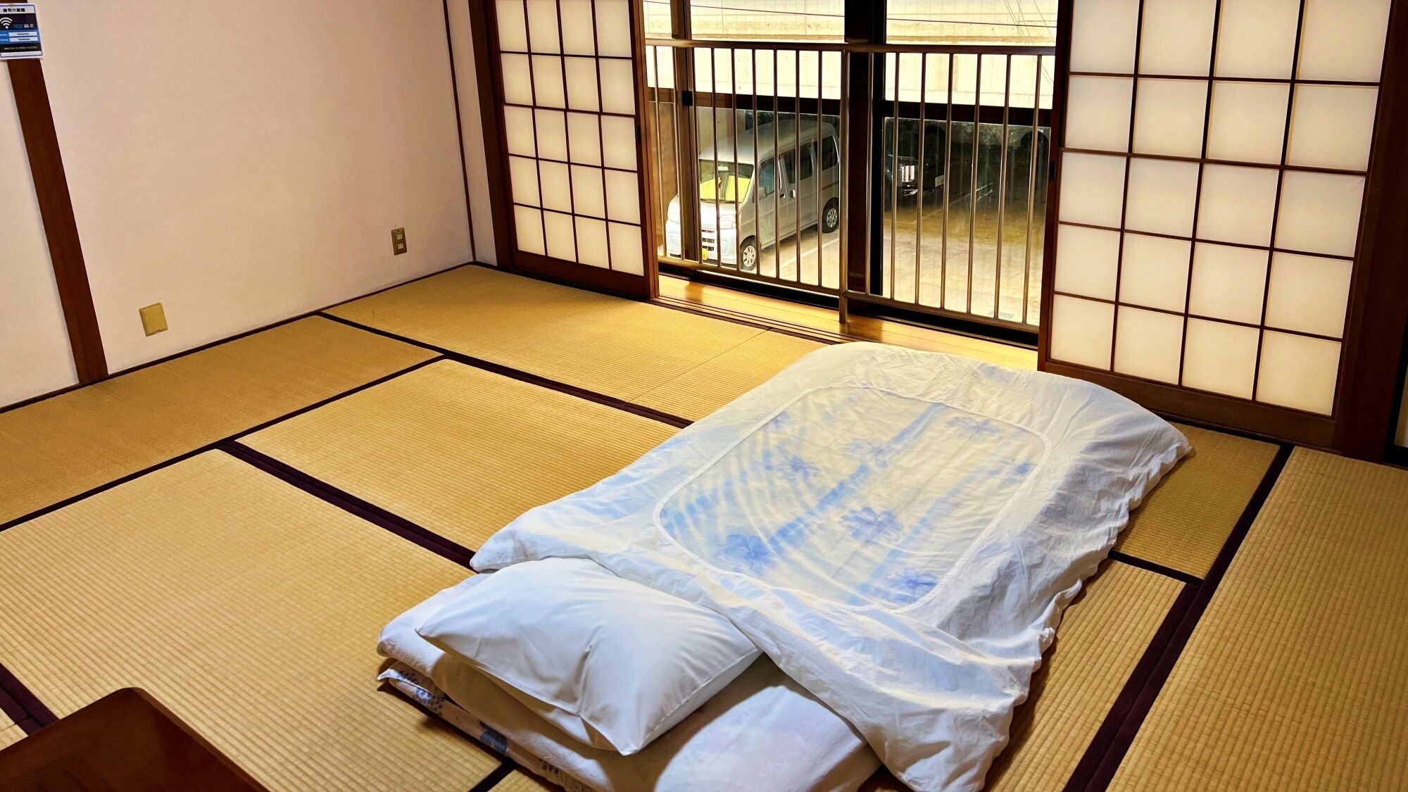 A tatami mat room with a bed, a sliding door, and a view of a parking area.