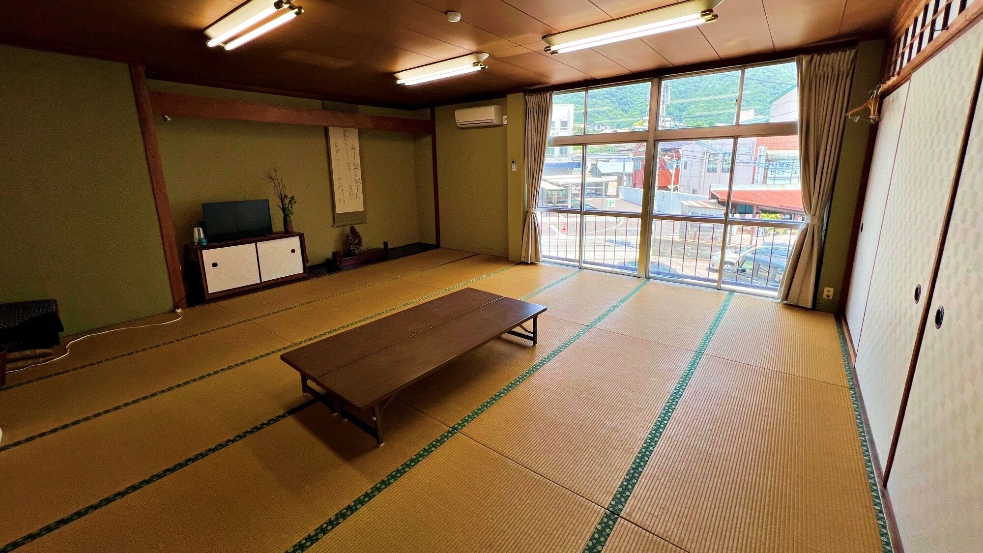 A tatami mat room with a wooden bench, a television, and a view of a town through large windows.
