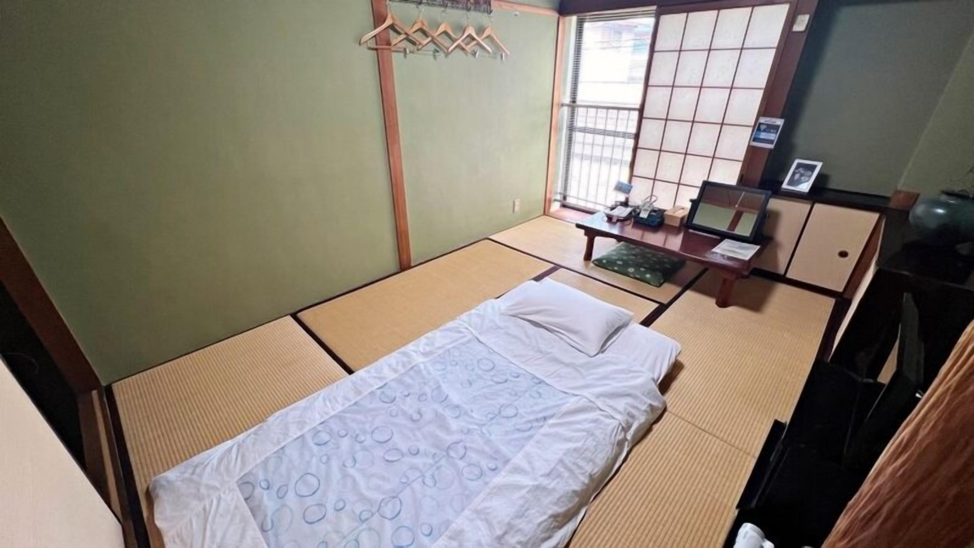 A room with tatami flooring, a bed with a white bedspread, a wooden desk with a chair, and a window with shoji screens.