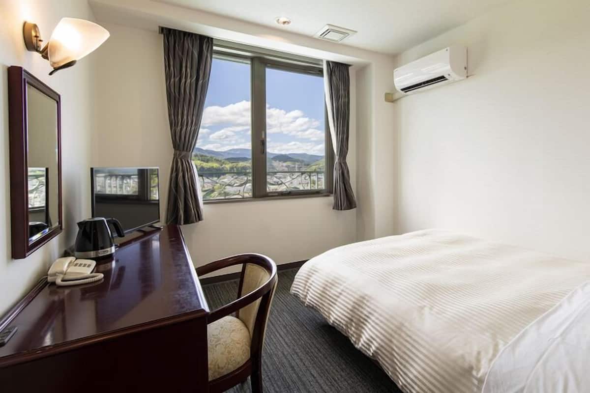 A hotel room with a bed, a desk with a telephone, a chair, a window with curtains, and an air conditioning unit.