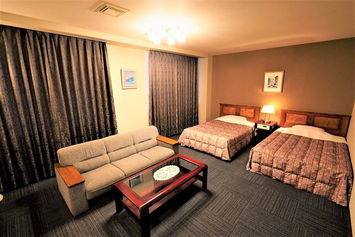 A hotel room with a sofa, two single beds, a nightstand with a lamp, and a small table with a decorative item.
