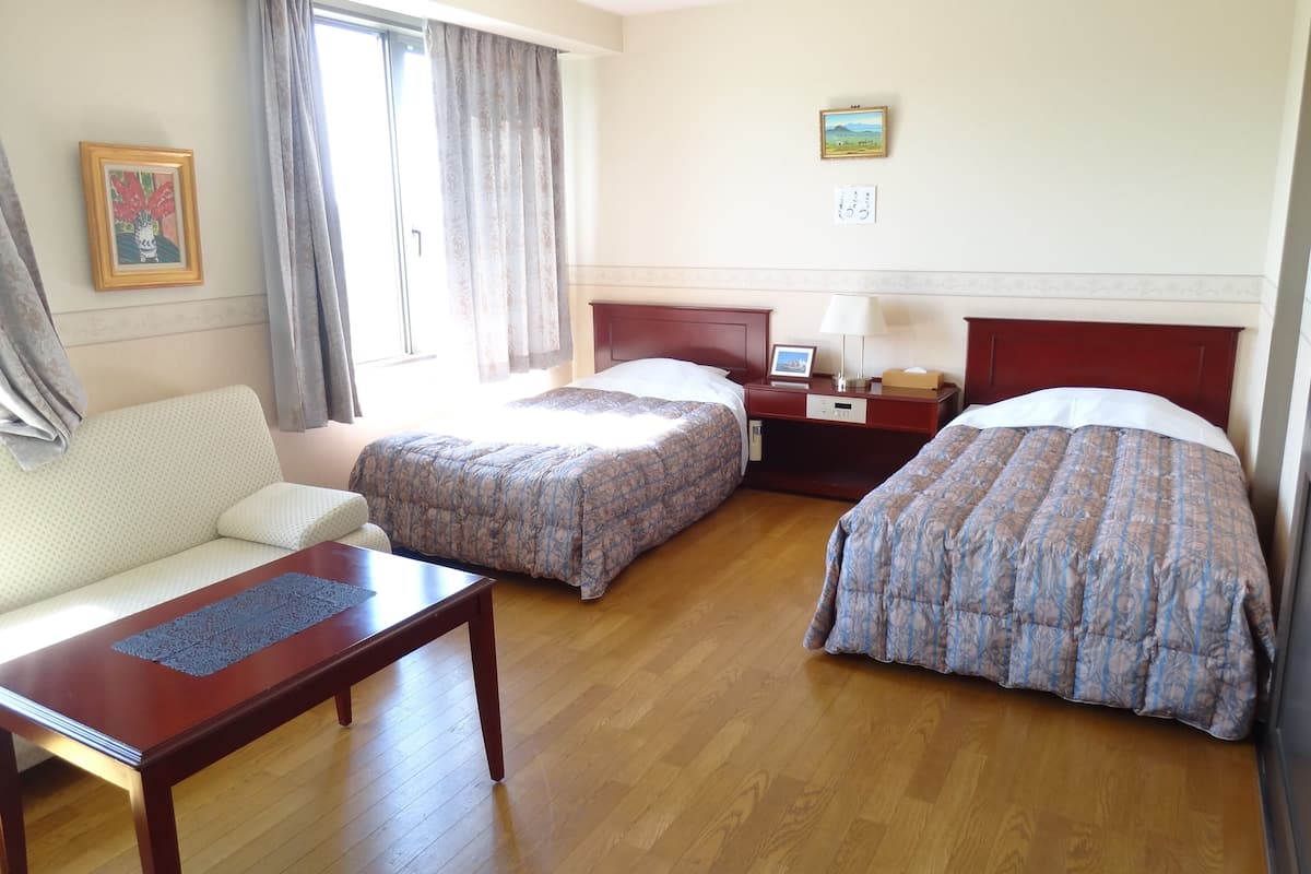 A hotel room with two beds, a sofa, a wooden table, a nightstand, and a window with curtains.