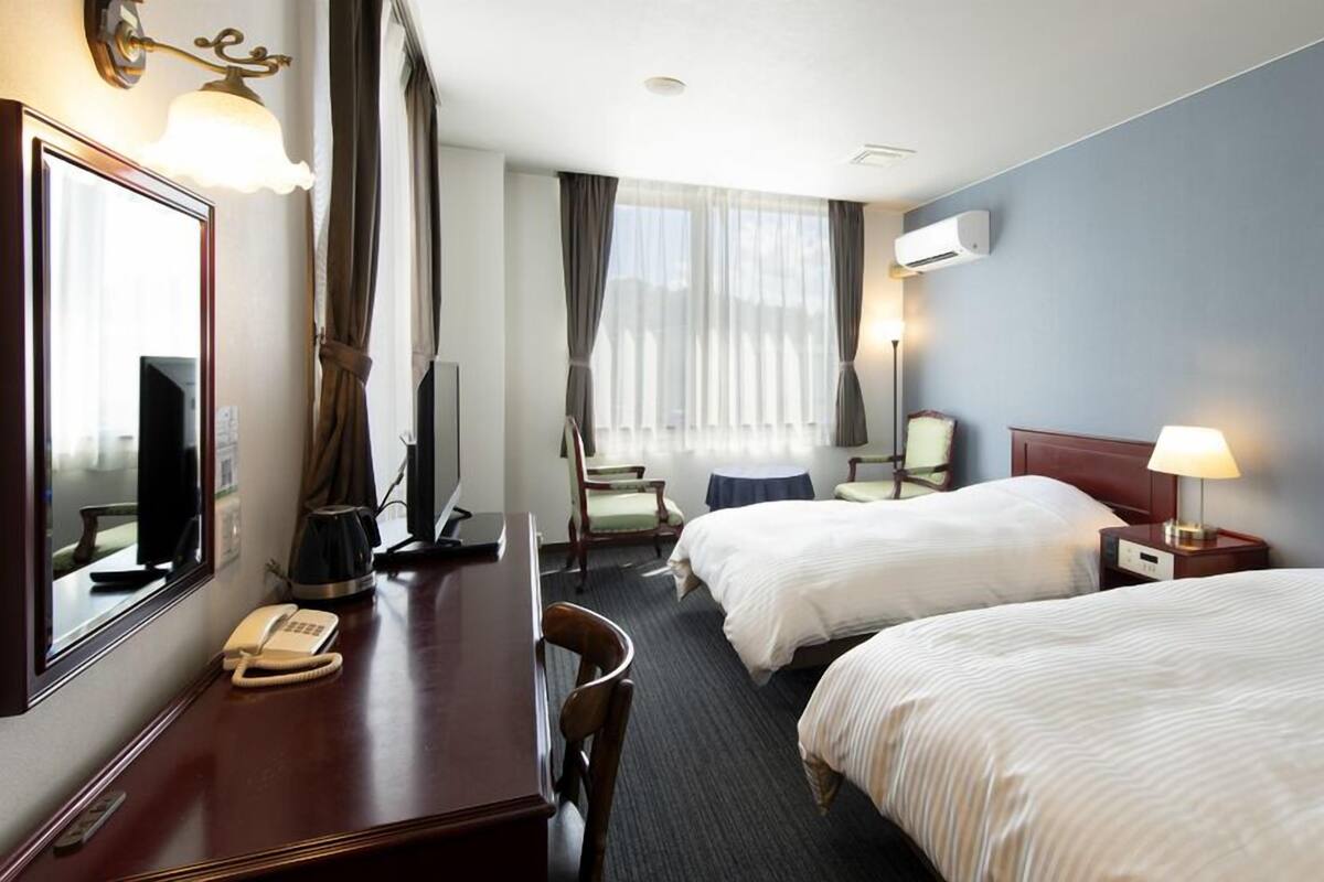 A hotel room with two beds, a desk with a telephone, a chair, a TV, and a window with curtains.