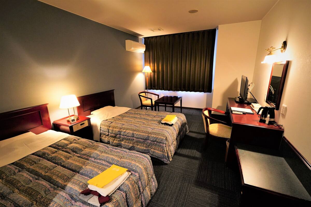A hotel room with two beds, a desk, a chair, a desk lamp, a mirror, and a window with curtains.