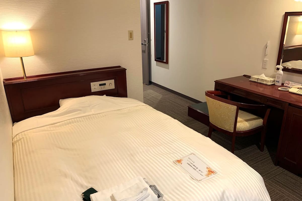 A hotel room with a bed, bedside lamp, desk, chair, and a mirror.