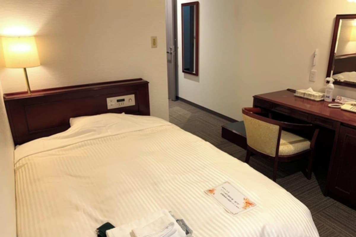 A hotel room with a bed, a desk, a chair, and a lamp.