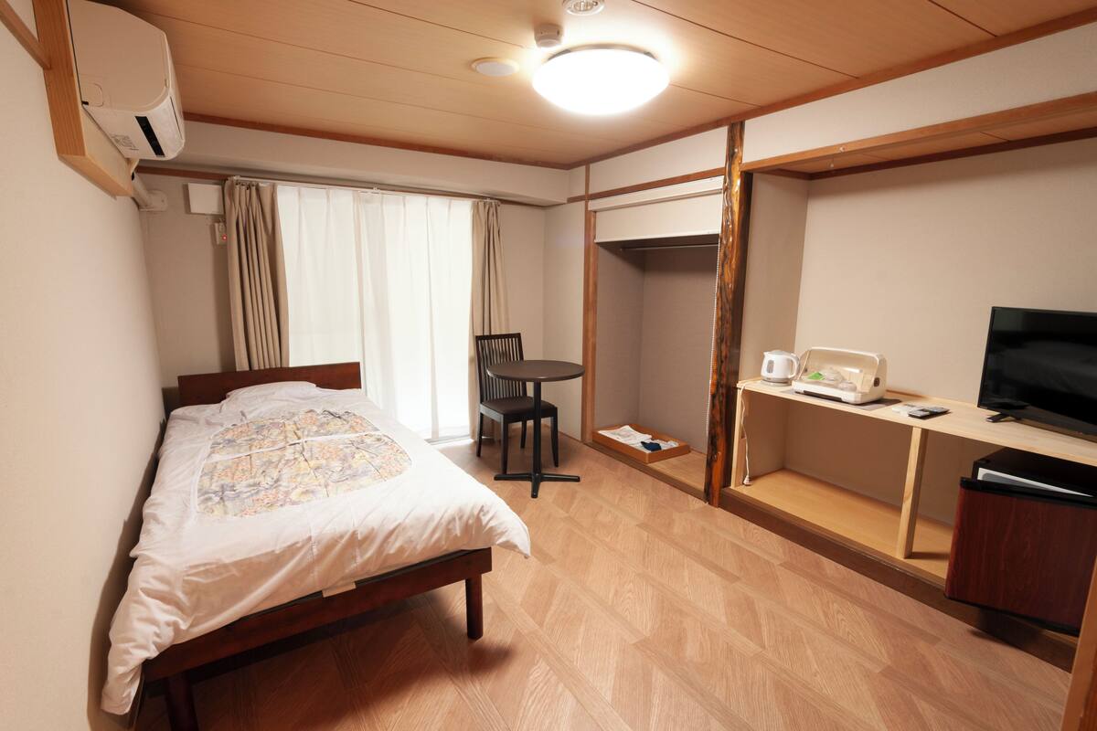 A hotel room with a bed, a desk with a TV, a chair, a table, and a wardrobe.
