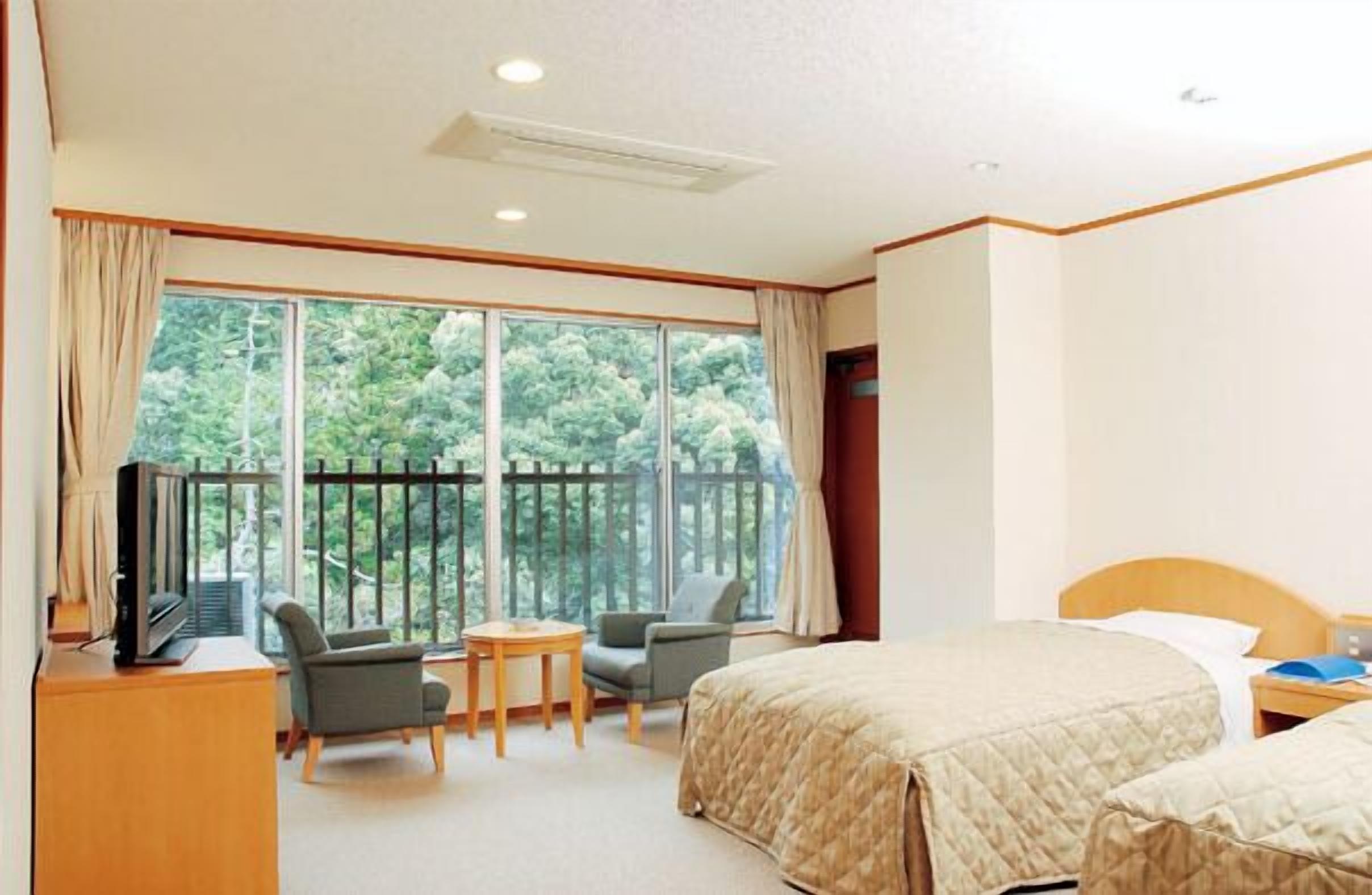 A hotel room with a large window, a bed, a desk with a TV, and two chairs.