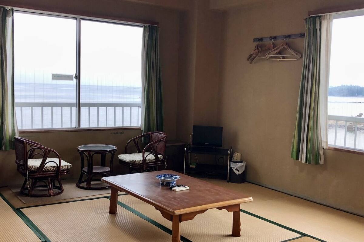 A room with tatami flooring, a low table, two chairs, a TV, and a view of a body of water through large windows.