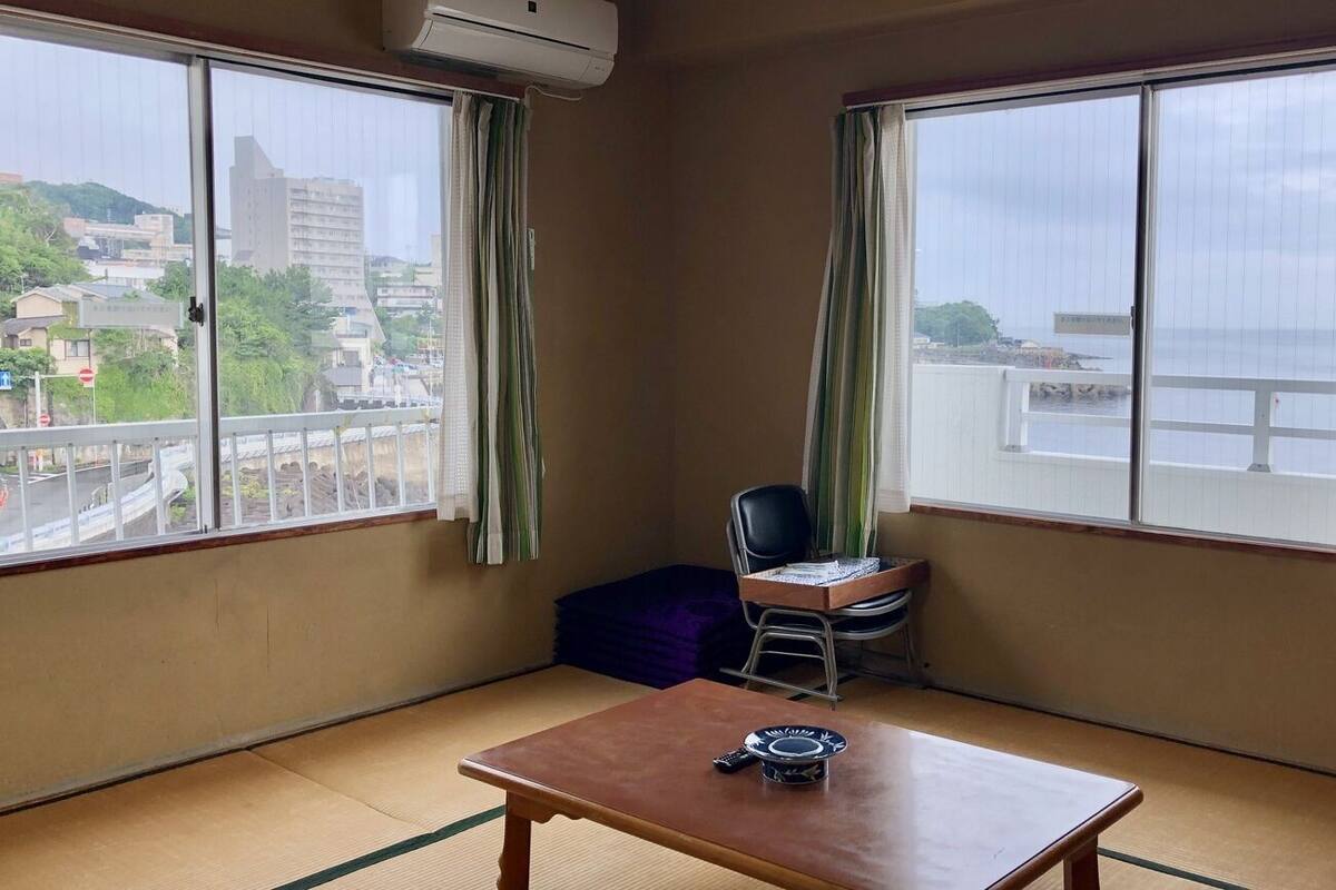 A room with tatami flooring, a wooden table, a chair, and a view of a cityscape through large windows.
