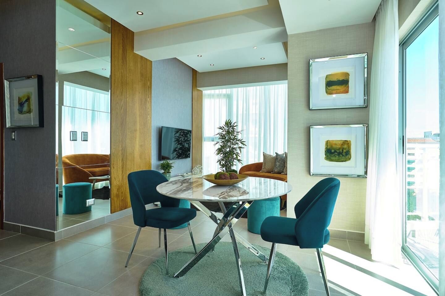 Un salon moderne avec une table ronde en verre, des fauteuils turquoise, un canapé et une grande fenêtre donnant sur la ville.