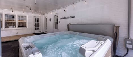 A hot tub with a sign reading "HOT TUB TIME POOL TOWELS" in a room with white brick walls, a ceiling fan, and string lights.