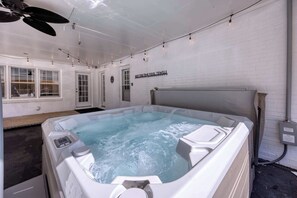 A hot tub with a sign reading "HOT TUB TIME POOL TOWELS" in a room with white brick walls, a ceiling fan, and string lights.