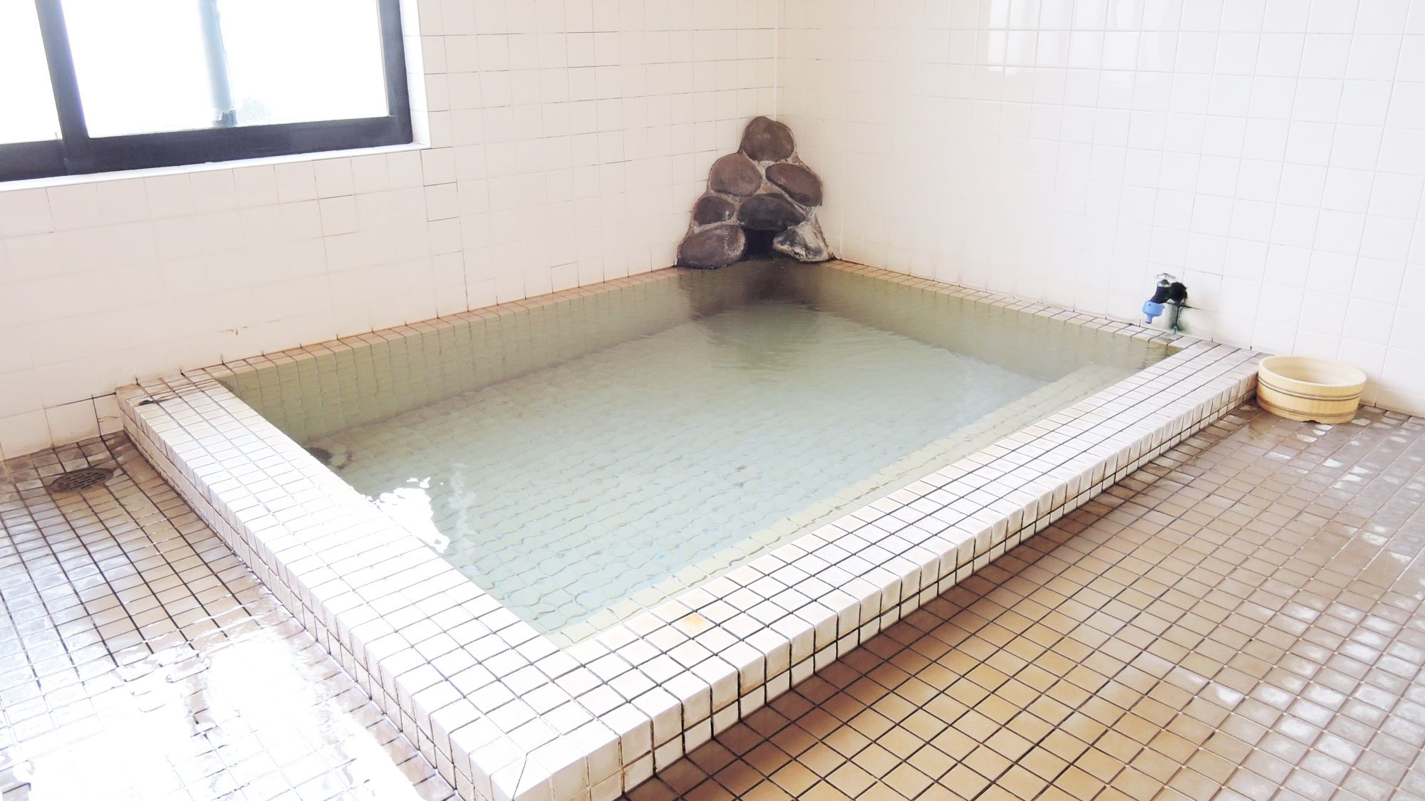 A rectangular hot tub with a stone structure in the corner, tiled walls, and a tiled floor.