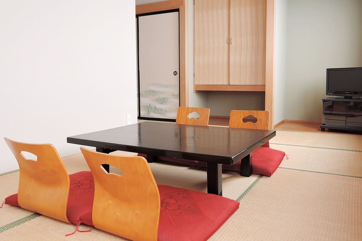 Quarto com piso de tatame, uma mesa baixa com cadeiras, televisão em um suporte e uma porta deslizante.