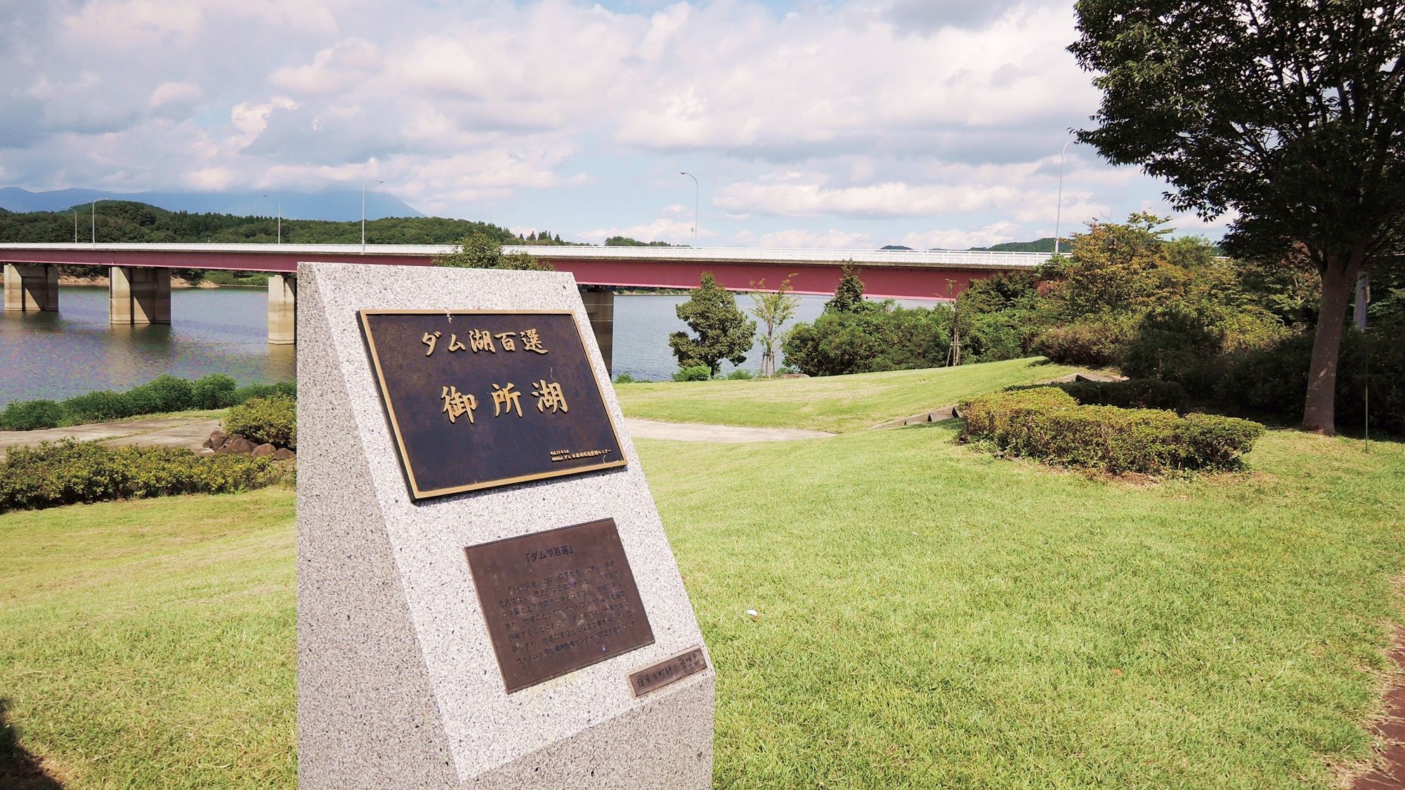 A stone monument with inscriptions, a grassy area, a bridge, and a body of water in the background.