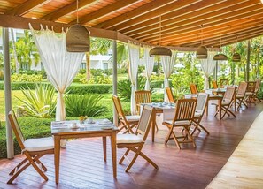 Een terras met houten tafels en stoelen, omgeven door groen en een houten pergola.
