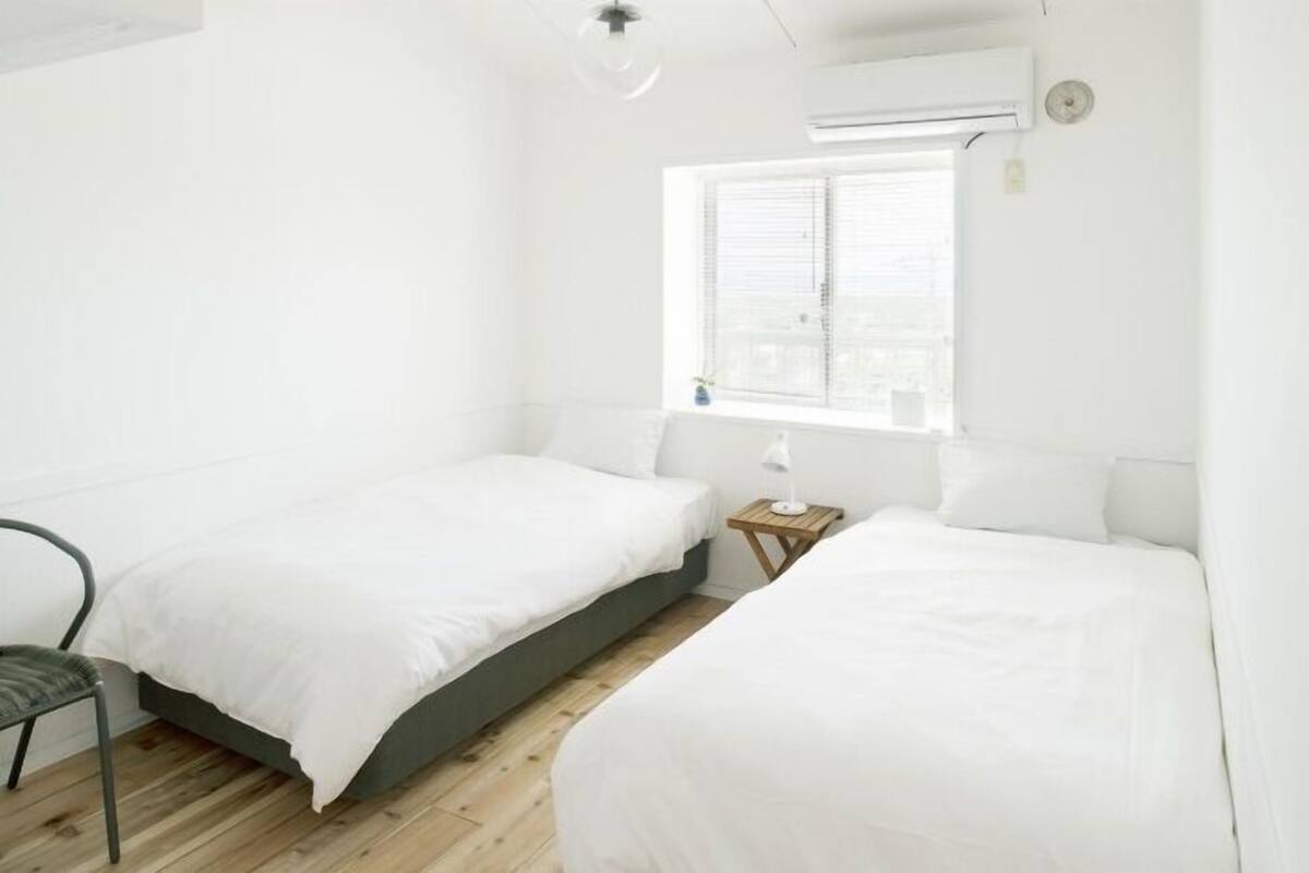 Una habitación con dos camas, una ventana con persianas, una mesita de noche con lámpara y una silla.