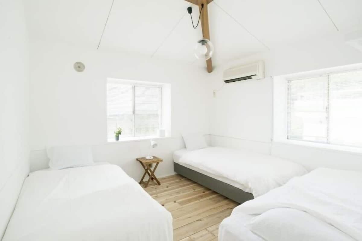 Una habitación con tres camas blancas, una pequeña mesa de madera, una ventana y un equipo de aire acondicionado.