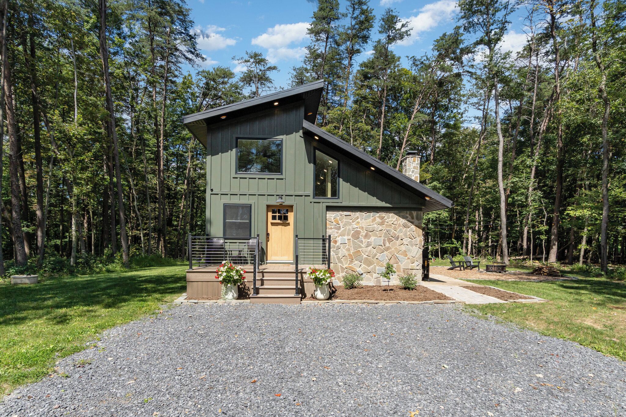 Une cabane à deux étages avec un mur en pierre, une terrasse en bois et une allée gravillonnée.