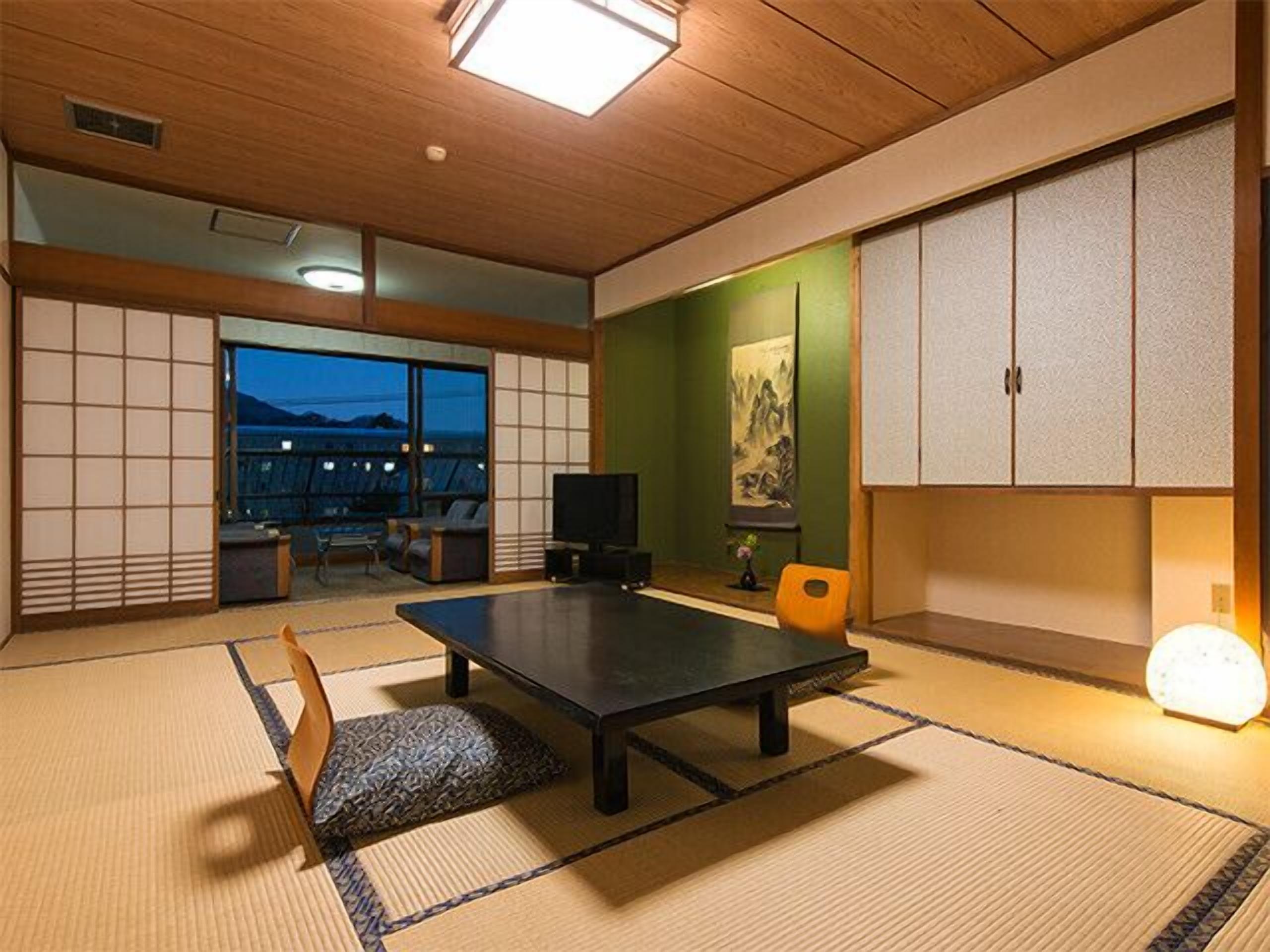 A traditional Japanese room with tatami flooring, a low table, a television, and a view of the mountains through large windows.