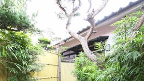 Un sendero de piedra que conduce a una puerta tradicional japonesa, rodeado de bambú y un edificio con techo de tejas.