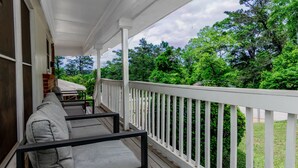 Eine überdachte Veranda mit weißer Reling, grau gepolsterten Sitzgelegenheiten und Blick auf üppiges Grün.