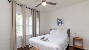 Ein Schlafzimmer mit Bett, Deckenventilator, Nachttisch und einem Fenster mit Vorhängen.