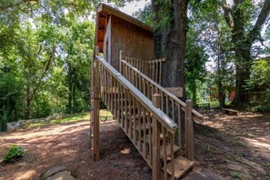 A wooden treehouse with a staircase, surrounded by dense trees. - Large Yard with Treehouse! Tri-Level Retreat (Columbus)