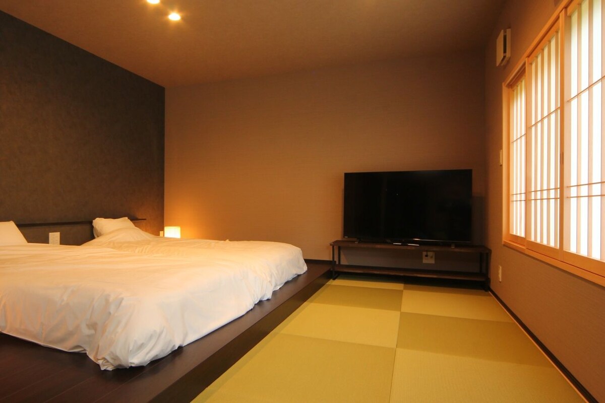 Ein Hotelzimmer mit Bett, Fernseher, Nachttischlampe und einem Fenster mit vertikalen Jalousien.