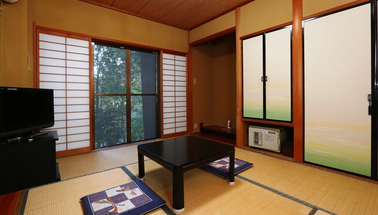 Ein traditioneller japanischer Raum mit Tatami-Böden, einem Fernseher, einem niedrigen Tisch, Schiebetüren und Blick auf Bäume.