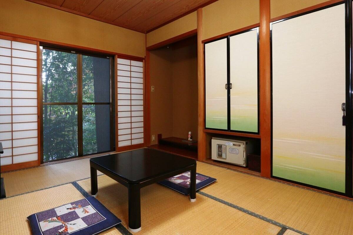 A traditional Japanese room with tatami flooring, a television, a low table, sliding doors, and a view of trees.