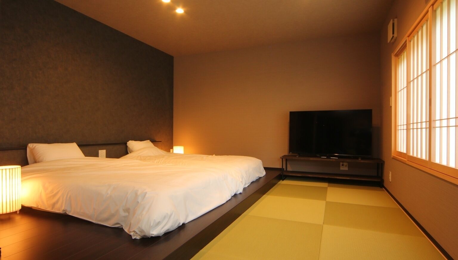 Ein Hotelzimmer mit Bett, Flachbildfernseher und Fenster mit vertikaler Jalousie.