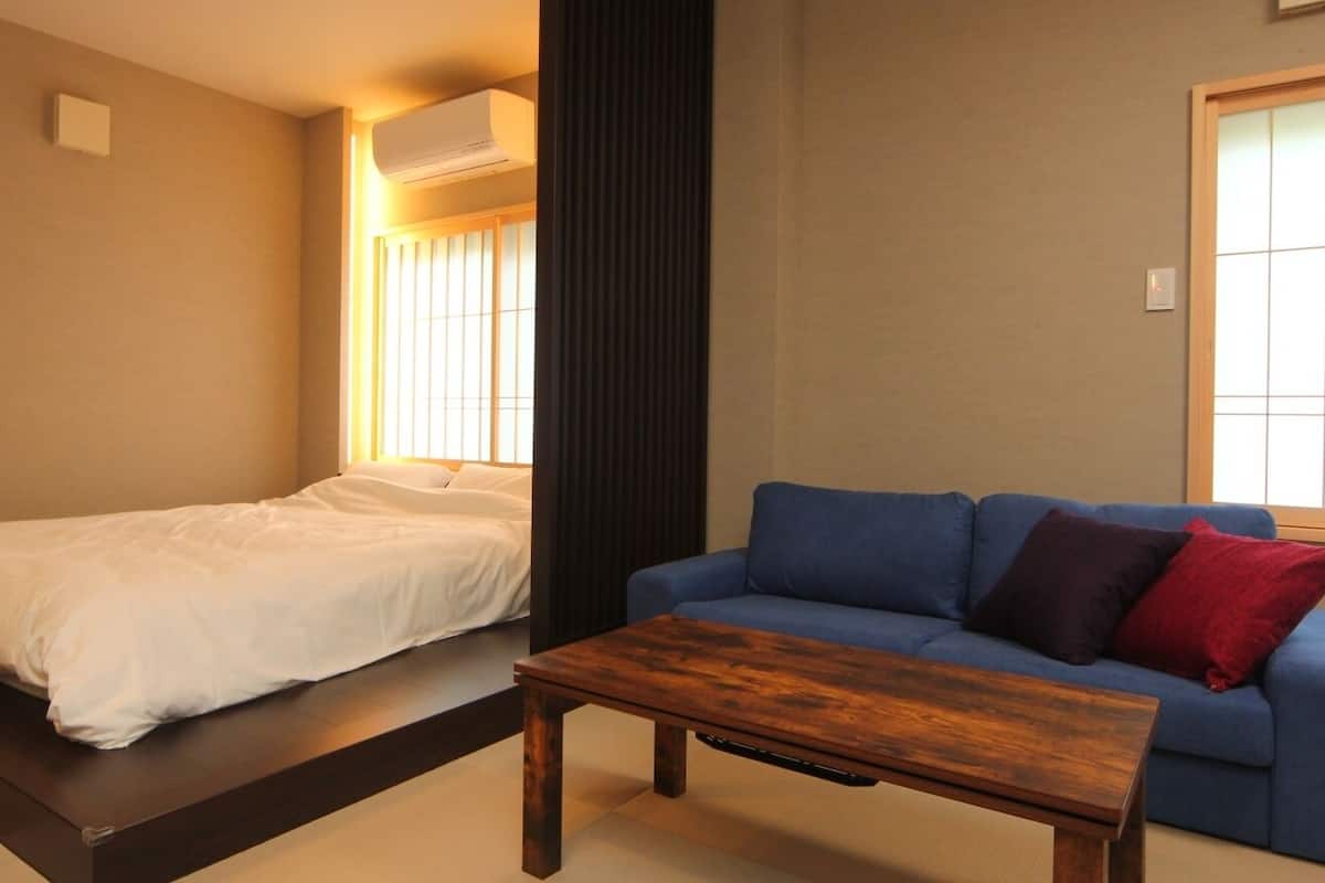 A room with a bed, a blue sofa, a wooden coffee table, and a window with blinds.