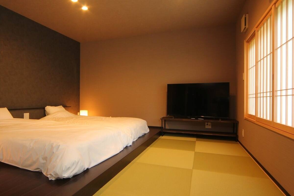 A hotel room with a bed, a TV, a bedside lamp, and a window with vertical blinds.