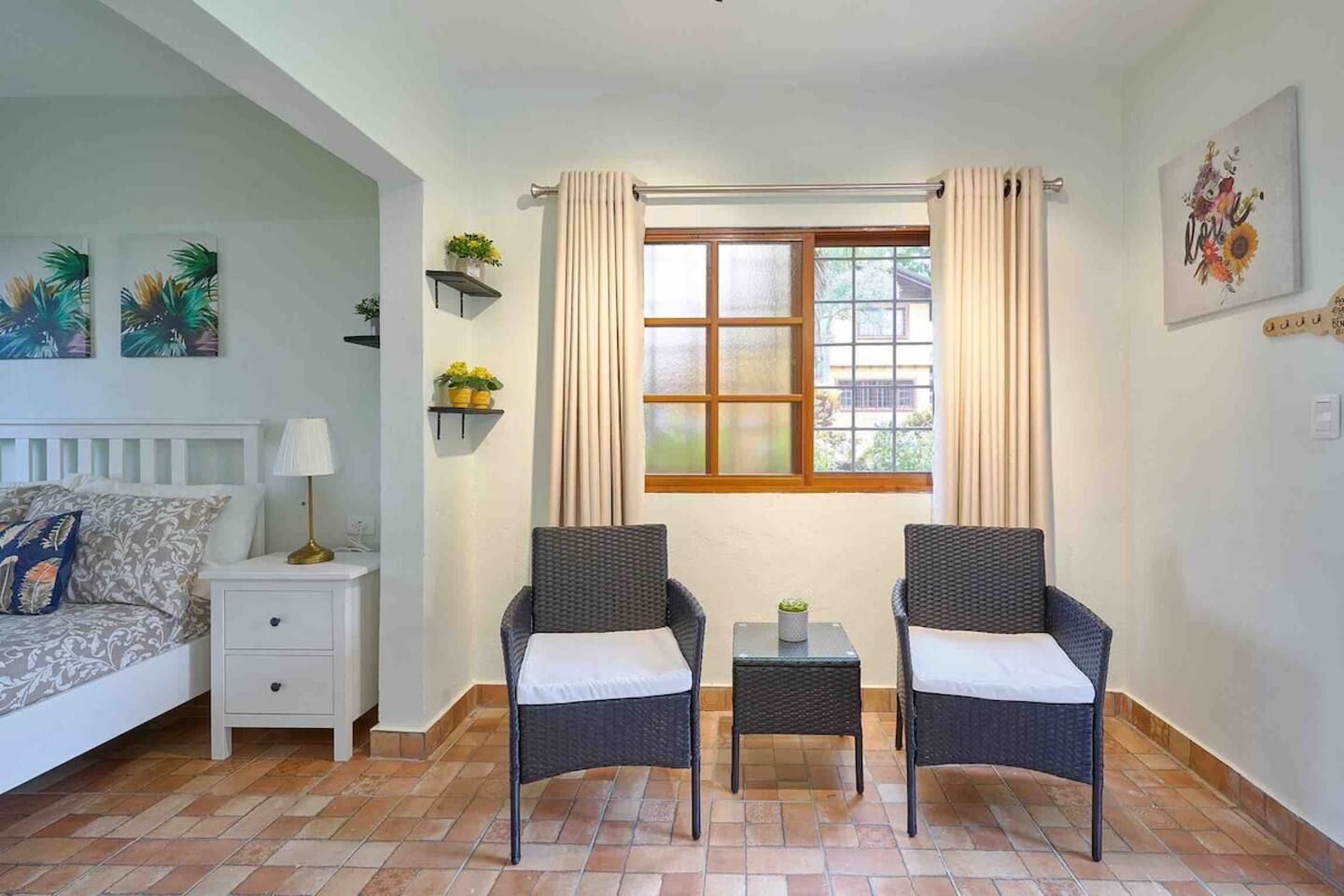 A room with a bed, two chairs, a small table, a nightstand, a window with curtains, and wall art.
