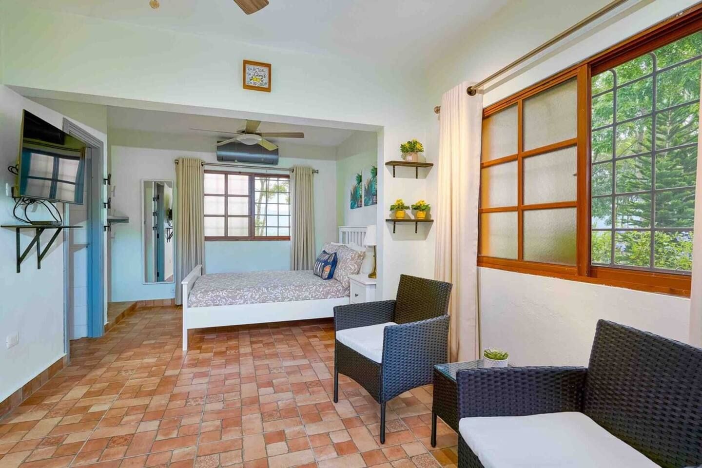 Een kamer met een bed, twee stoelen, een plafondventilator, een raam met gordijnen en een klein plankje met planten.
