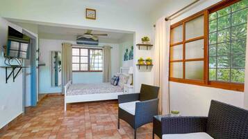 A room with a bed, two chairs, a ceiling fan, a window with curtains, and a small shelf with plants.