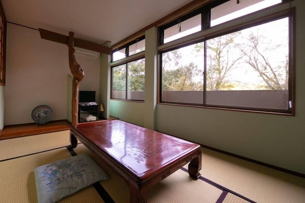 A tatami mat room with a wooden table, a pillow, a shelf with items, and large windows.