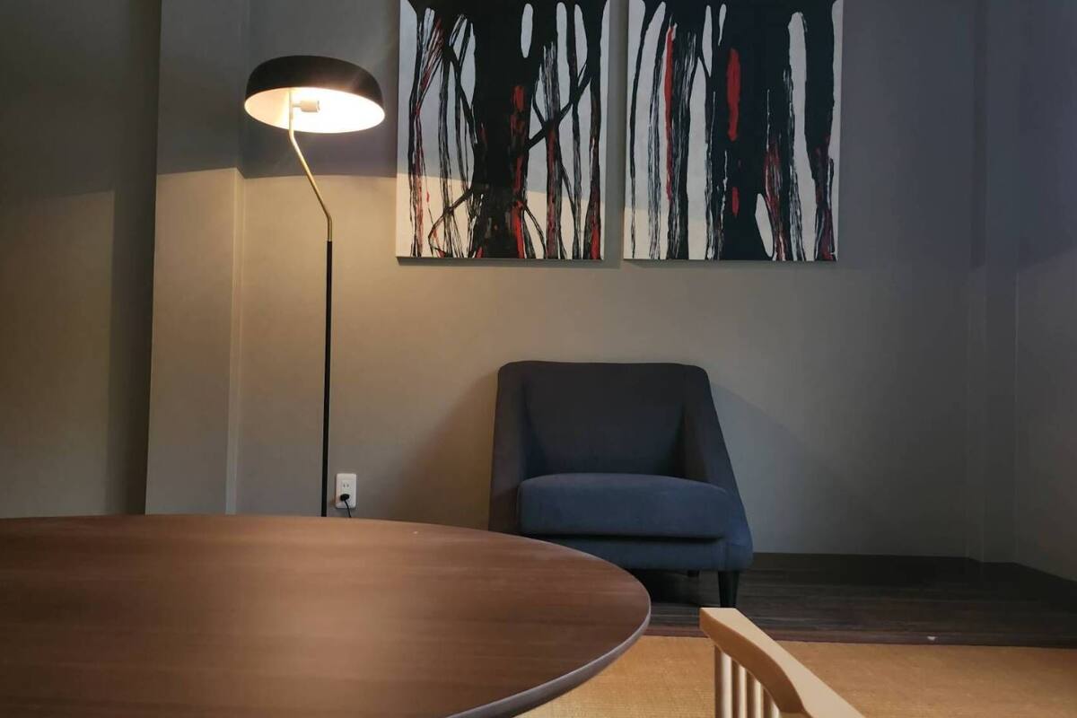 A room with a round wooden table, a blue chair, a floor lamp, and a large abstract painting on the wall.