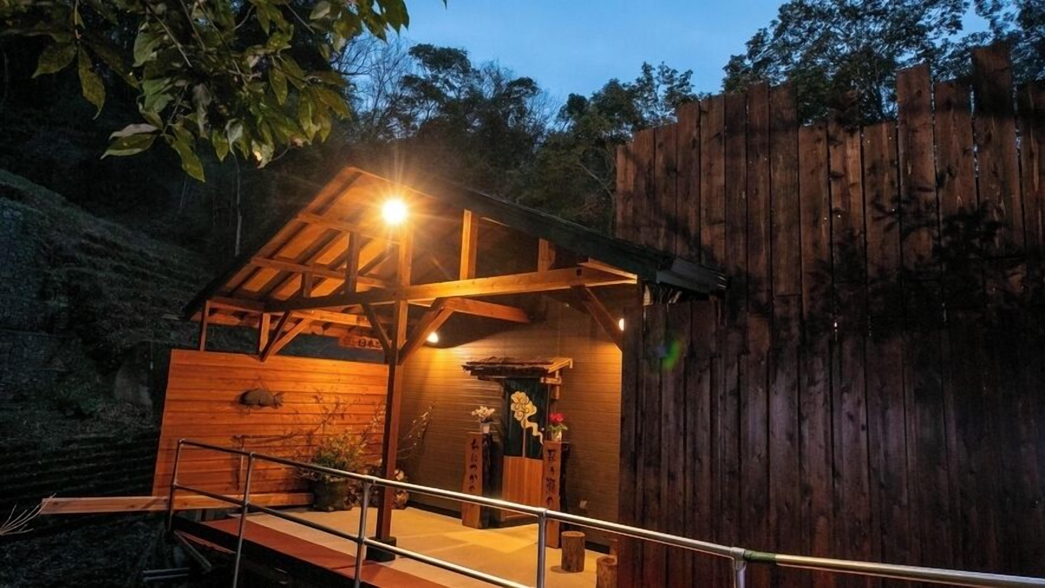 A wooden cabin with a porch, a small shrine, and a wooden fence in the background.