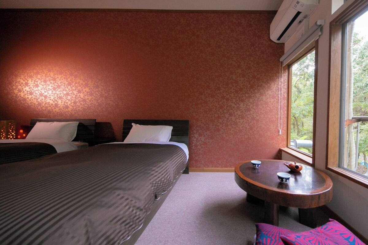 A hotel room with a bed, a small round table with a teapot, a window with a view of trees, and an air conditioning unit.