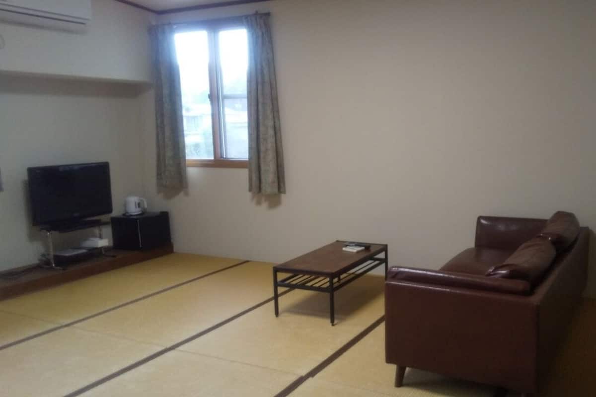 A living room with tatami flooring, a sofa, a coffee table, a TV, and a window with curtains.